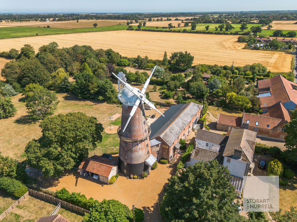 Aerial view of a restored brick windmill and long barn surrounded by trees and harvested fields